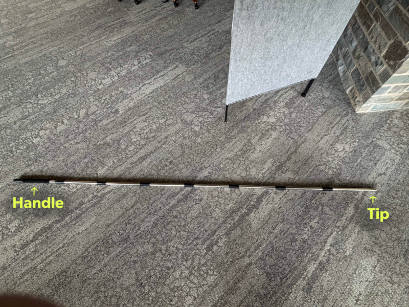 Step One A long extendable pole assembled and placed on a gray carpeted floor. Bright yellow text on the image points out the tip and the handle of the pole.
