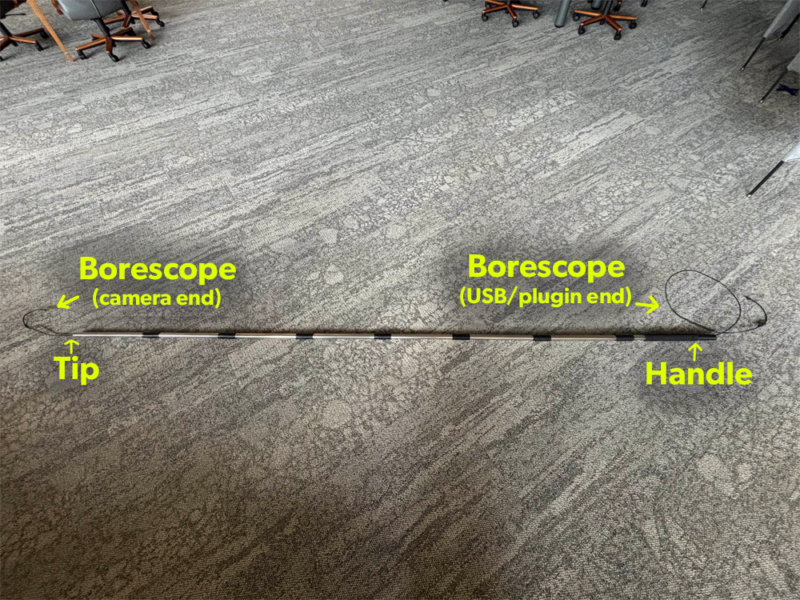 A long extendable pole assembled and placed on a gray carpeted floor. bright yellow text on the image points out the tip and the handle of the pole, as well as each end where the borescope appears. The camera end of the borescope emerges from the tip of the pole, and the USB/plugin side of the borescope wire emerges from the handle end.