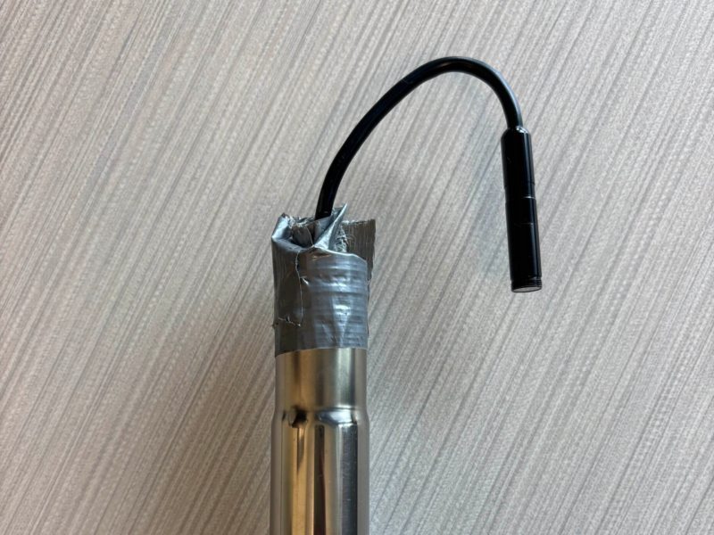 Step Three The top of a metal pole with duct tape attaching the end of an endoscope that has been threaded through the hollow pole. The tip of the endoscope has been bent in a U-shape to look back down towards the rest of the pole, like a candy-cane.